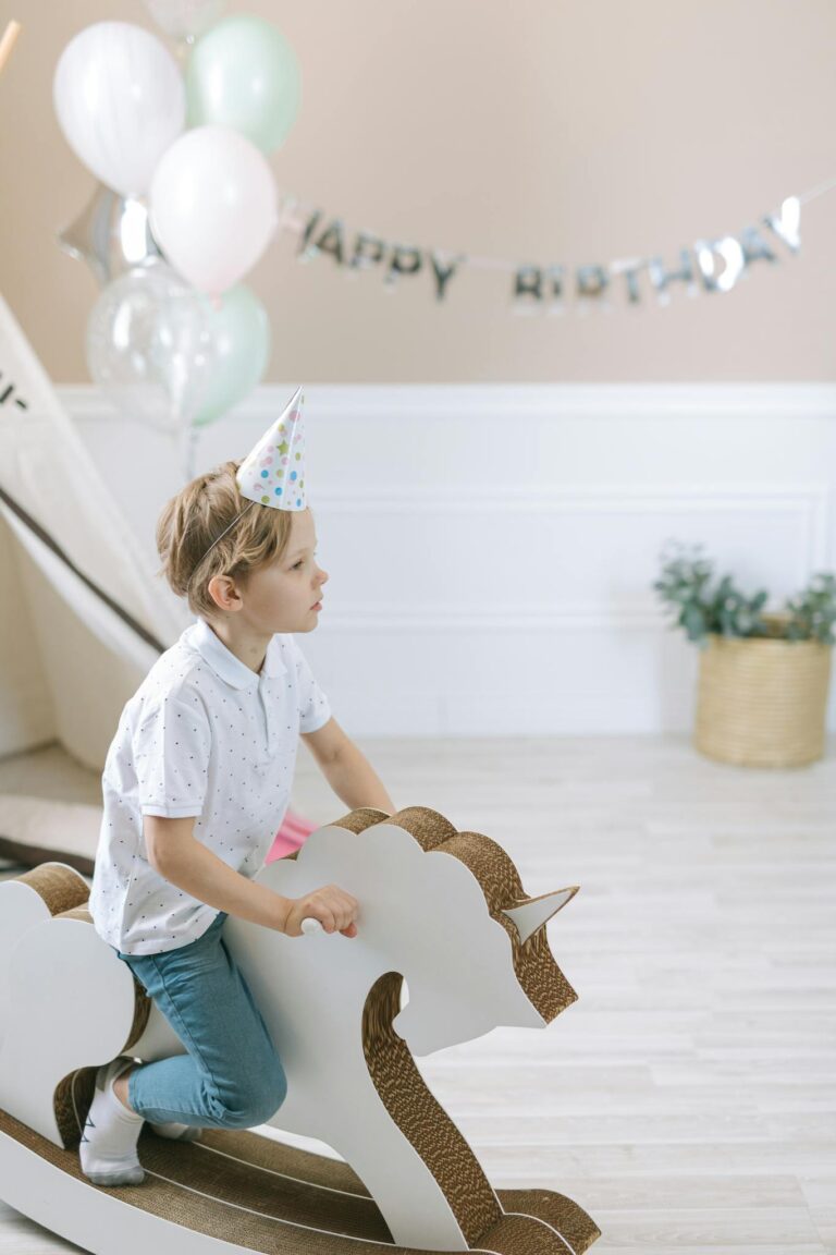 A young boy with a party hat rides a wooden unicorn rocking horse at a birthday celebration.