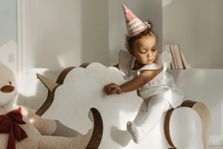 A young girl in a birthday hat sits on a rocking unicorn toy indoors.